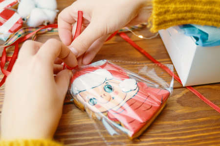 Close up of cookies with colored icing on wooden table. Christmas gifts. Womens hands tie red ribbon on bag of gingerbread in form of pig in Santa Claus hat. New Years atmosphere.の写真素材