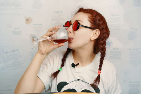 Cheerful female in round red goggles with funny pigtails drinks wine. Hippie woman holding glass of...
