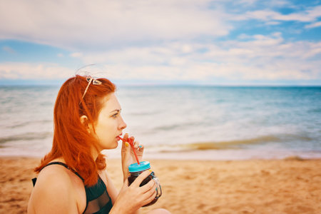 Red-haired girl in a swimsuit is sitting on the beach with a cocktail. Calm woman on vacation. Holidays at the sea. Vintage sunglasses on the head of a having a rest lady.の写真素材