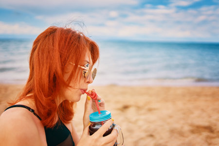 Red-haired woman with sunglasses on vacation. Holidays at the sea. Calm lady in a swimsuit is sitting on beach with a cocktail and look away. Tropical alcohol beverage. Summer day with cloudy sky.の写真素材