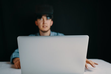 A young man with a VR headset is focused on using a laptop in a dimly lit room, showcasing his hightech engagementの写真素材