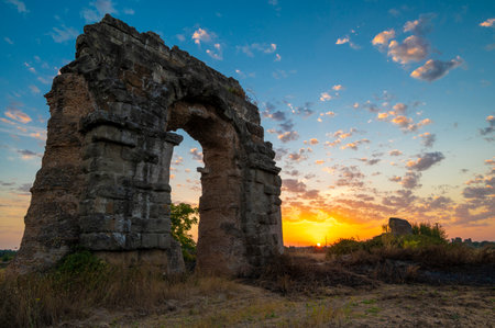 Park of the aqueducts ruins of the Claudius aqueduct, Via Appia Antica. Perspective arch during sunset, play of clouds, blue sky, yellow-orange sun reflections on the horizon. Rome, Italy. Appio latino districtの写真素材