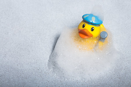 A yellow rubber duck for a bath in a bath cap and with a shower head in the wing in a cloud of soapy foamの写真素材