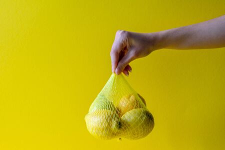 Fresh yellow lemons in plastic nettingin a female hand on a yellow background . Food background textureの写真素材