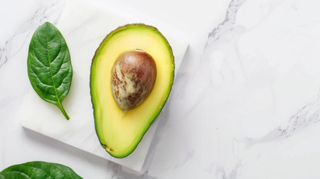 A fresh sliced avocado reveals its creamy texture and seed, positioned on a marble surface alongside a vibrant green leaf, symbolizing healthy living.の素材