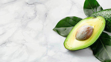A fresh halved avocado with a seed sits on a smooth marble surface, surrounded by vibrant green leaves, symbolizing healthy eating and natural nutrition.の素材