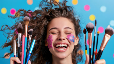 A joyful young woman exuberantly poses with a collection of makeup brushes, her face adorned with colorful paint against a vibrant dotted background, showcasing creativity and fun.の素材
