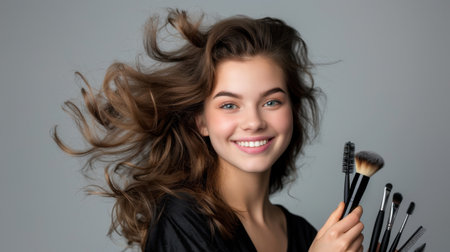 A joyful young woman with flowing hair displays her makeup brushes against a gray backdrop, radiating beauty and confidence in this lively portrait.の素材