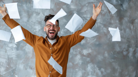 A cheerful man celebrates his success, throwing papers into the air with a radiant smile. The bright indoor space features a textured gray wall, adding a dynamic and lively atmosphere to the moment.の素材