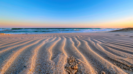A tranquil beach scene depicting soft sand patterns illuminated by the warm hues of sunrise. Gentle waves lap the shore, creating a soothing atmosphere.の素材