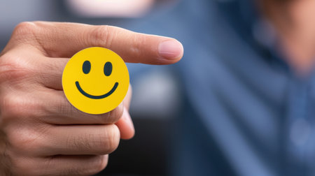 A hand shows a cheerful gesture while holding a bright yellow smiley sticker, symbolizing positivity and happiness in a vibrant office setting.の素材