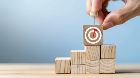 A close-up of a hand placing a wooden block with a target symbol on a stack of cubes, illustrating the concepts of goal achievement and strategic planning.の素材