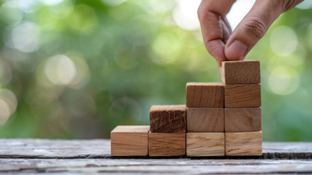 A hand carefully places wooden blocks in a stacked formation, illustrating the theme of growth and progress against a soft, blurred natural backdrop.の素材
