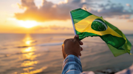 A stunning view of a person holding the Brazil flag against a breathtaking sunset over the ocean. The golden sky reflects on calm waters, creating a serene atmosphere.の素材
