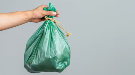 A person's hand holds a tied green plastic garbage bag filled with waste, symbolizing efforts in waste management and promoting environmental sustainability.の素材