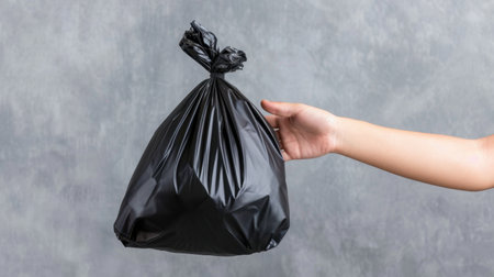 A hand holds a tied black plastic garbage bag, symbolizing waste disposal and recycling efforts. Ideal for concepts of cleanliness and sustainability.の素材