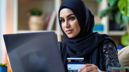 A focused young woman in a hijab engages in online shopping using a credit card at her laptop in a cozy home office, displaying modern lifestyle and technology.の素材