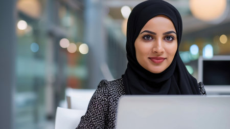 A confident woman in a hijab works intently on her laptop in a stylish office environment, showcasing focus and professionalism in a contemporary workspace.の素材