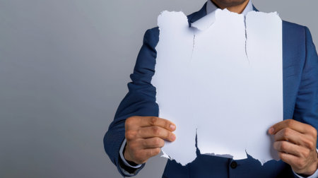 A businessman in a formal suit stands against a gray background, holding a torn piece of paper. The image conveys themes of rejection and disappointment, ideal for various business contexts.の素材