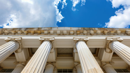 A stunning low-angle view of grand architectural columns reaching up toward a bright blue sky adorned with fluffy white clouds. Ideal for showcasing classic design.の素材