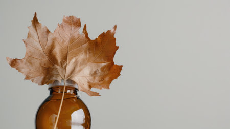 A stunning brown glass bottle topped with a dried maple leaf, perfect for autumn-themed decor, showcasing a warm and creative aesthetic in a neutral setting.の素材