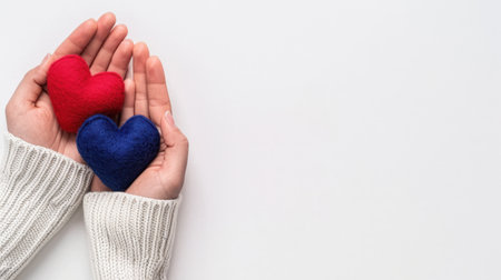 Two vibrant felt hearts, one red and one blue, are gently held in hands against a minimalist white background, conveying love and care.の素材