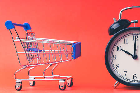 An empty shopping cart and an alarm clock on a red scarlet background. Shopping, online shopping concept, time to buy concept, copy space, top view,の写真素材