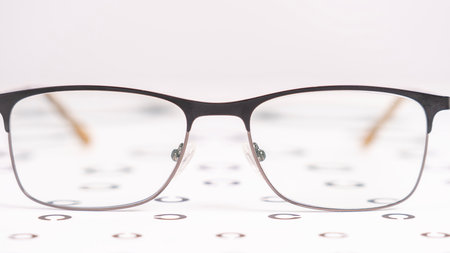 A pair of glasses is on a table with a white background. The glasses are black and have a gold frame