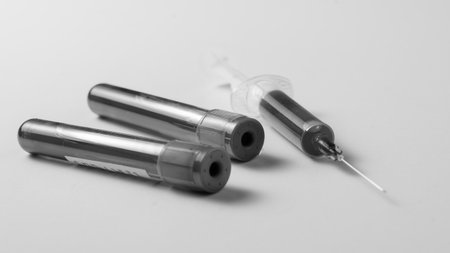 A syringe and two blood tubes are placed on a white surface. The syringe is positioned between the...