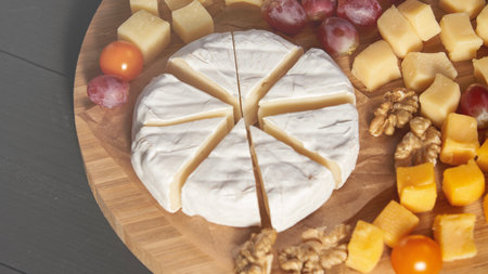 A wooden board with a wedge of cheese and a variety of fruits and nuts. The cheese is cut into six...