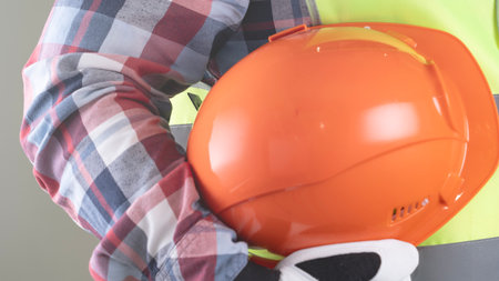 A person wearing a yellow and orange safety vest is holding an orange hard hat. The person is wearing a plaid shirt and is holding the hard hat in their right handの写真素材