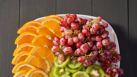 A plate of fruit with oranges, kiwis, and grapes. The plate is on a wooden tableの写真素材