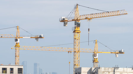 Three yellow cranes are in the sky, one of which is taller than the others. The cranes are in a city, and the sky is cloudyの写真素材