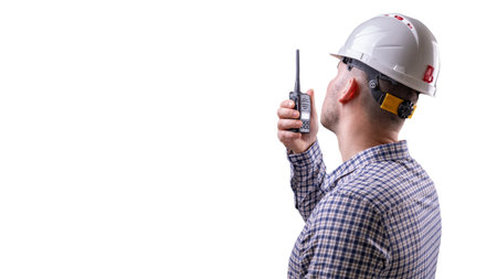 A construction worker, wearing protective gear, transmits instructions over the radio, while observing the progress of the work on the site. He is isolated on a white background and looks into the distance. High quality photoの写真素材