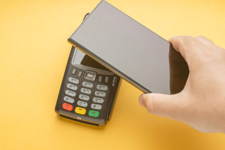 Hand holds phone near payment terminal. Yellow background adds brightness to scene. Action suggests...