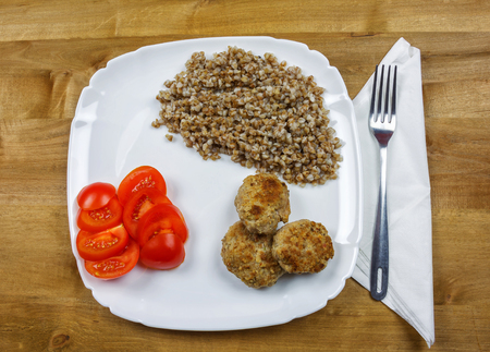 On a square plate is buckwheat porridge with meat cutlets and chopped tomatoの写真素材