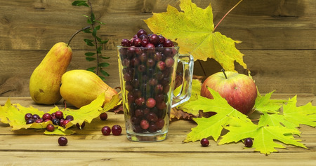 Against the background of the wooden surface is a glass beaker with cranberries, autumn leaves lie side by side, an apple and a pear.の写真素材