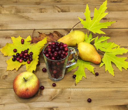 On a wooden surface is a glass beaker with cranberries. Next to them are autumn leaves, two pears and an apple.の写真素材