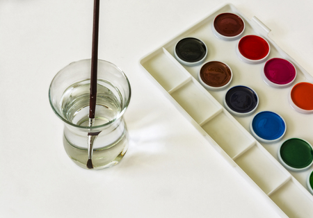 On a white surface is a set of watercolor colors and there is a glass with water and a brushの写真素材