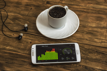 On the wooden surface there is a Cup of black coffee on a saucer, next are headphones and a smartphone with an equalizerの写真素材
