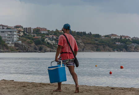 Sozopol, Bulgaria - 09/06/2018: a man in a red t-shirt with a blue box walks along the beach. Stock photoのeditorial素材