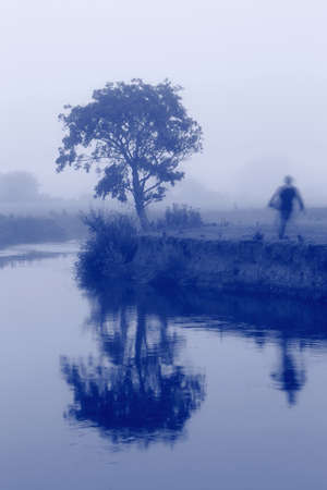 Tree reflected on the river Axe in Devon with blurred person passing by. Monochrome blue tone imageの写真素材