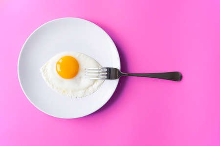 Breakfast, fried egg, yolk, omelet and fork on white plate with color table, creative pictureの写真素材