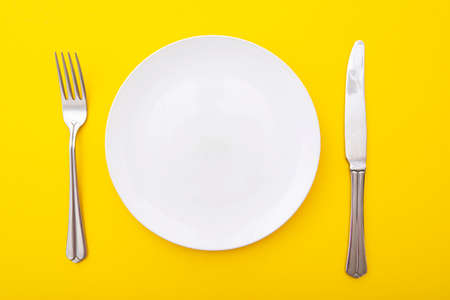 Ceramic white plate with knife and fork on yellow surface, kitchen table, lay out, top view, space...