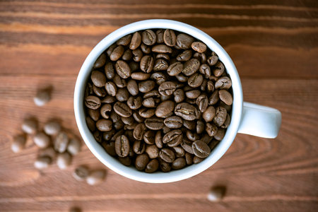 Heap of coffee beans in white ceramic mug on wooden table. Roasted coffee grains on wooden surface,...