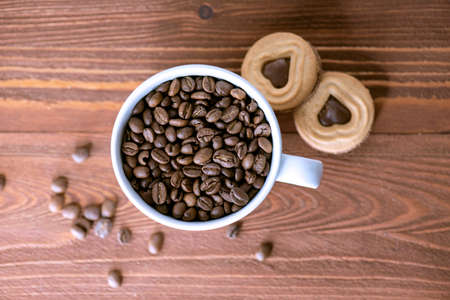 Coffee grains or beans in a ceramic mug with biscuits on wooden table. Top view of roasted coffee seeds.の写真素材