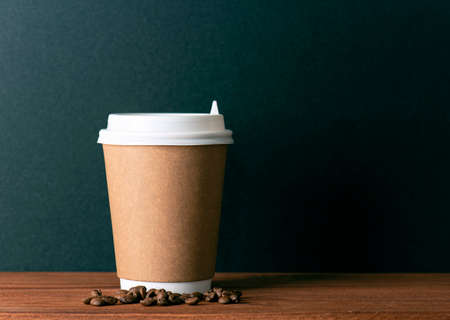 Disposable paper cup with hot coffee and roasted coffee grains on wooden table in front of black wall. Space for textの写真素材