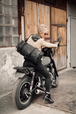 Woman sitting on her motorcycle. Motorcycle near the garage. A woman in a leather jacket. Motorcycle black. A woman with a short haircut. Bikeの写真素材