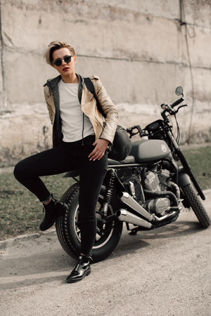 Woman sitting on his motorcycle. Motorcycle near a grey wall. girl with short hair. girl in black glasses and a leather jacket. Bikeの写真素材