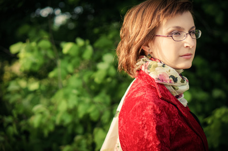 Portrait of a young beautiful woman posing outdoors in a red velvet coat and a scarf with bright flower pattern.の写真素材